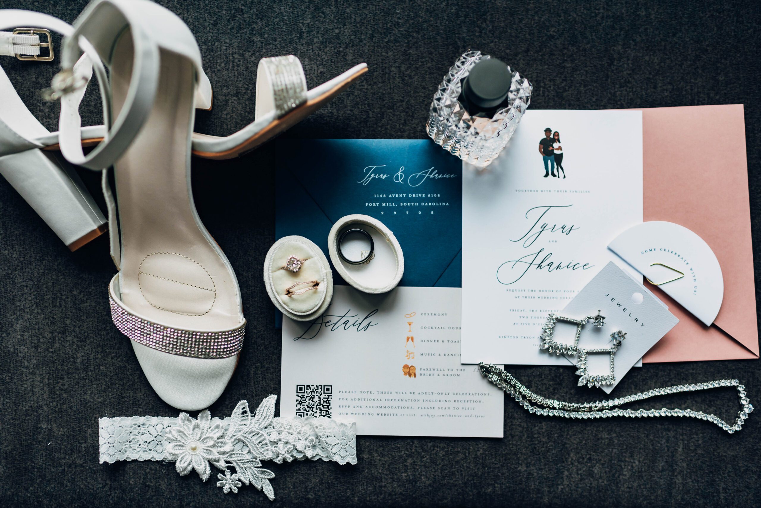 Wedding flat lay including perfume, shoes, wedding invitations with caricature, rings, garter, necklace, earrings, and envelope